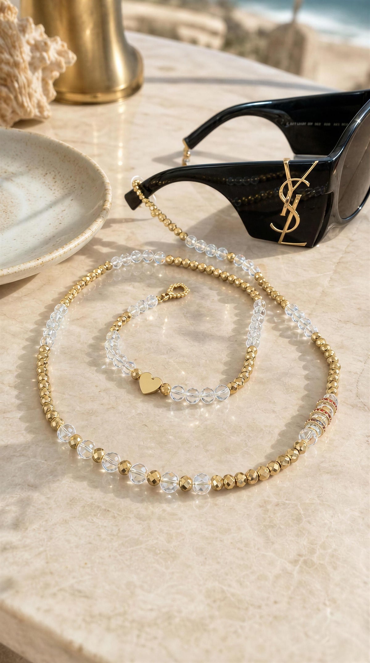 The Riviera Sunglass Chain - Cristalle features alternating clear and gold beads with a delicate heart charm, elegantly draped next to black designer sunglasses on a marble surface.
